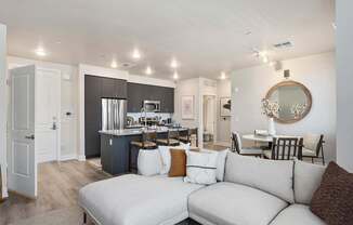 A spacious open living area here at Reflect at Dobson Ranch featuring a light neutral sectional with textured accent pillows, wood-style flooring, and recessed lighting, flowing into a modern kitchen with dark cabinetry, an island with bar seating, and stainless-steel appliances, alongside a cozy dining nook with a round table, wall mirror, and soft decorative accents.