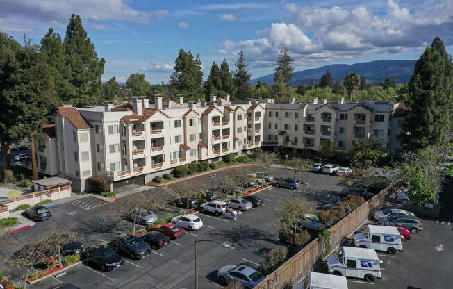 Aerial view of a multi-story residential building surrounded by trees and a parking lot filled with cars. The sky is partly cloudy, and mountains are visible in the background.