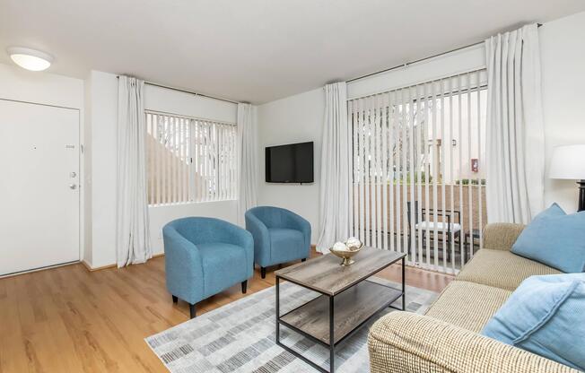 A cozy living room featuring a beige sofa with blue cushions, two blue armchairs, a modern coffee table, and a flat-screen TV mounted on the wall. Large windows with vertical blinds allow natural light to enter. The floor is wooden, and there's a decorative bowl on the coffee table, creating an inviting atmosphere.