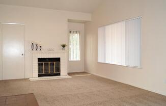A light-colored living room featuring a fireplace with a white mantel, beige carpeting, and a large window with vertical blinds. The walls are bare, creating a minimalist appearance, and a door is visible on the left side of the room.