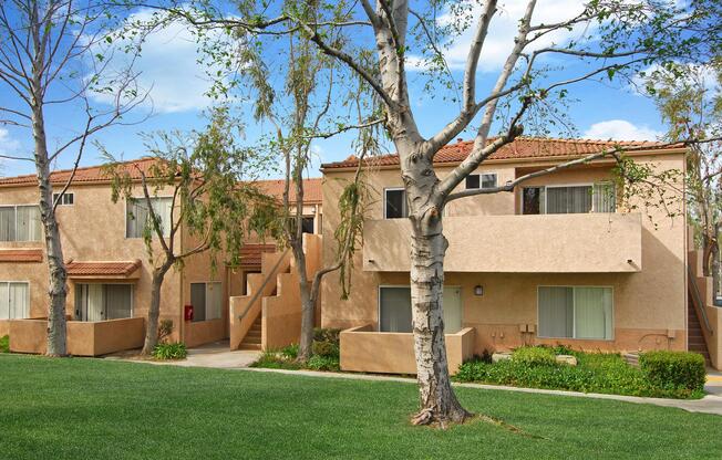 A peaceful residential scene featuring beige two-story apartments surrounded by greenery. The buildings have balconies and large windows, with steps leading to the upper levels. Leafless trees add to the serene atmosphere against a blue sky with fluffy clouds. Perfect for a tranquil living environment.