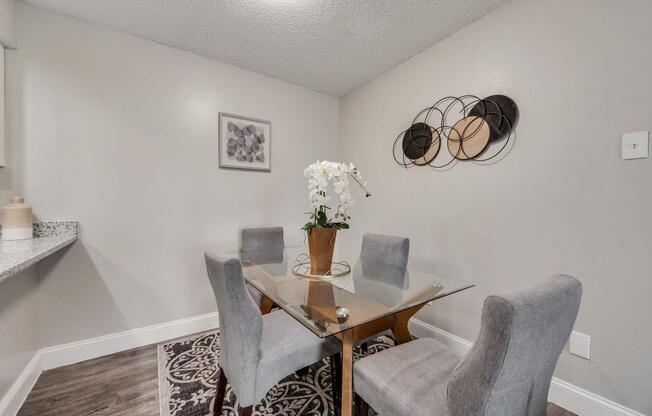 A modern dining area featuring a glass table surrounded by four gray upholstered chairs. A potted white orchid centerpiece sits on the table. The walls are a soft gray, adorned with a decorative wall hanging and a piece of artwork. A patterned rug adds warmth to the space.