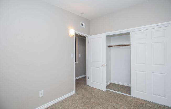 Interior view of a light-colored room featuring beige carpet, a closet with sliding doors, and a light fixture on the wall. One door leads to another room, while the closet door is open, revealing a wooden shelf inside. The walls are painted a soft gray, creating a serene atmosphere.