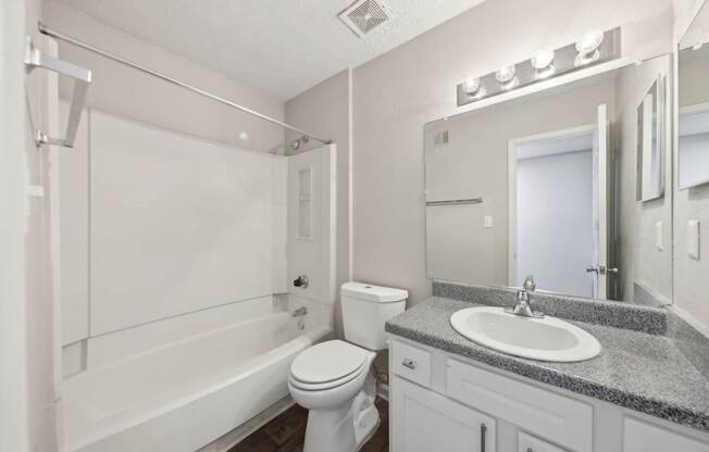A bathroom with a white bathtub and shower, toilet, sink, and a mirror with lights at Gwinnett Square Apartments in Duluth, GA