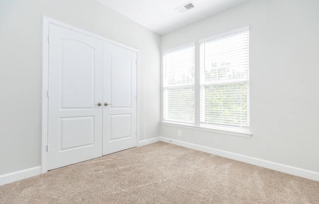 A bright, empty room with light gray walls and a tan carpet. There are two large windows with white blinds allowing natural light to fill the space. A double door closet is visible on one wall, adding to the room's simplicity and openness.