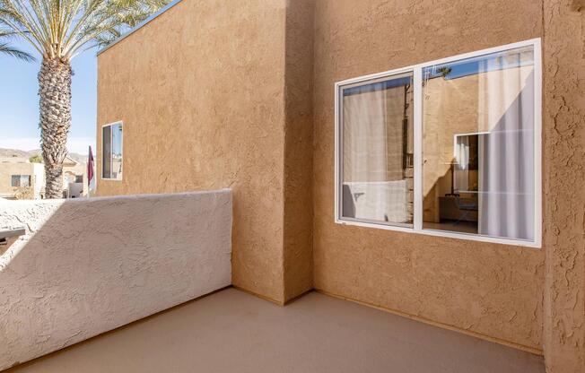 View of a sunlit outdoor patio area with textured beige walls and a large window reflecting the interior. A palm tree is visible in the background, adding a tropical feel to the scene. The floor is smooth and neutral-colored, suggesting a warm, inviting space.