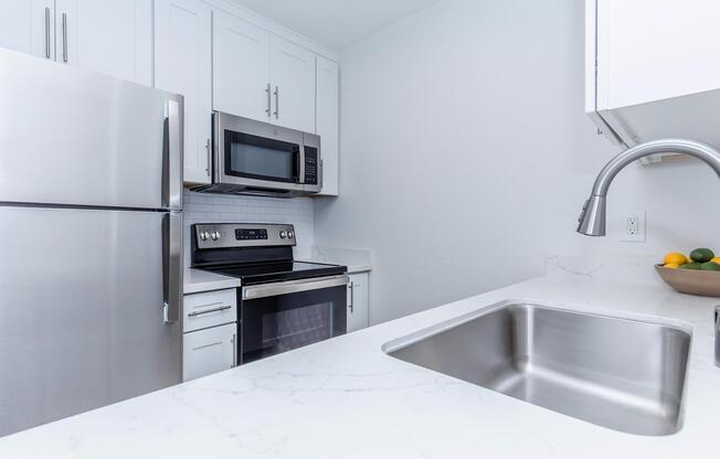 Modern kitchen featuring stainless steel appliances, including a refrigerator, microwave, and oven. The countertop is white with a marble-like finish, and a stainless steel sink is visible. Light-colored cabinets line the wall, contributing to a bright and clean aesthetic. A bowl of fruit is placed on the counter.