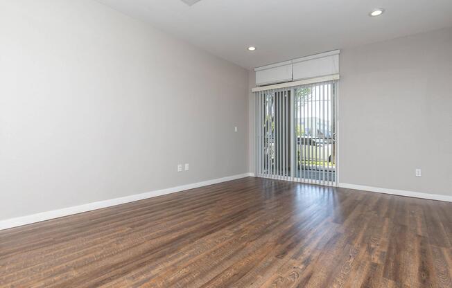 An empty room with light gray walls and wooden flooring. There is a sliding glass door with vertical blinds leading to an outdoor area. The space is well-lit with overhead lights, creating a bright and open atmosphere.