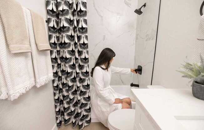 A woman in a white robe is sitting on a toilet in a bathroom.