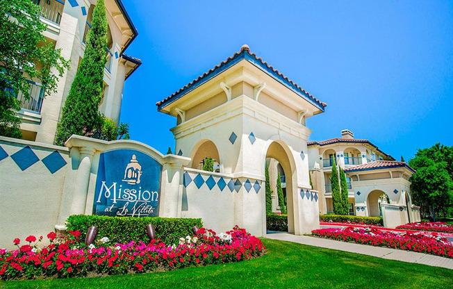 Community entrance with florals and luscious grass at Mission at La Villita Apartments in Irving, TX offers 1, 2 & 3 bedroom apartment homes with appliances.