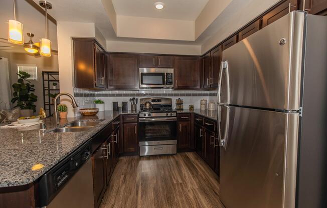A modern kitchen featuring dark wooden cabinetry, stainless steel appliances, and granite countertops. It includes a stove, microwave, and dishwasher, complemented by a sleek refrigerator. The space is well-lit with pendant lighting and has a small dining area with greenery on the counter.