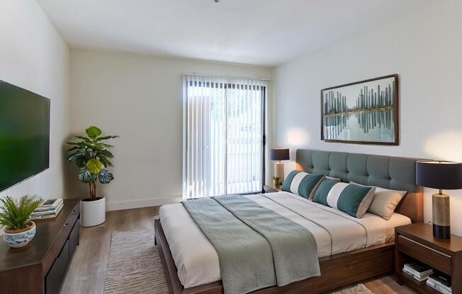 A modern bedroom featuring a neatly made bed with decorative pillows, a stylish headboard, and side tables with lamps. A large television is mounted on the wall, and a potted plant adds a touch of greenery. Natural light streams in through sliding glass doors, enhancing the serene atmosphere.