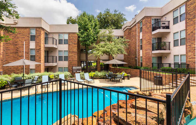 A swimming pool with a waterfall surrounded by lounge chairs and a couple of tables, chairs and umbrellas at Cambridge Court Apartments Entryway in Lake Highlands, Dallas, TX.