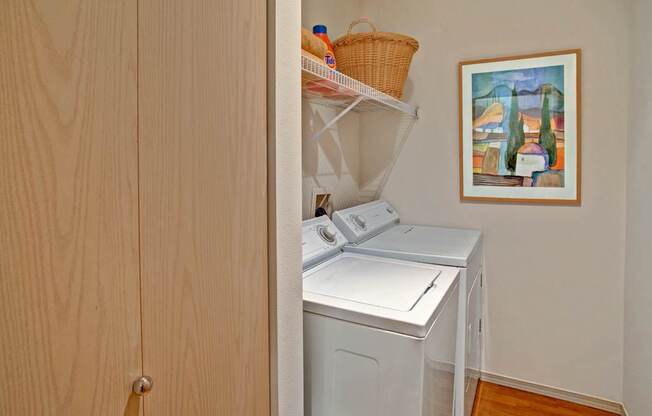A compact in-home laundry area here at Portsmith featuring a side-by-side washer and dryer set tucked into a recessed nook, white wire shelving above with storage baskets and supplies, light wood cabinet doors, warm wood-look flooring, and a framed wall art accent creating a clean and practical laundry space.