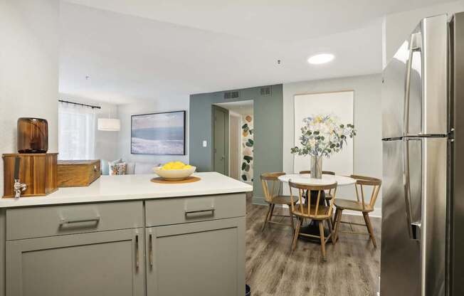 A kitchen with a refrigerator, a dining table with chairs, and a bowl of fruit on the counter.