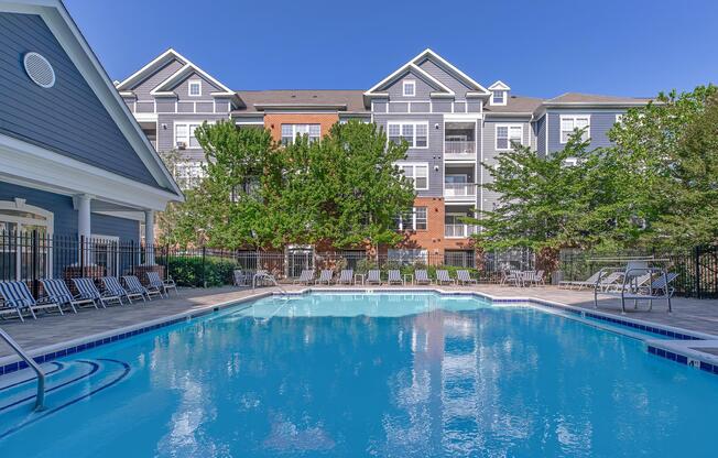 A clear swimming pool surrounded by lounge chairs, with a backdrop of a multi-story residential building. The scene is set under a blue sky and features green trees lining the pool area, creating a welcoming atmosphere for relaxation and leisure.
