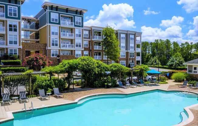 A swimming pool surrounded by lounge chairs and trees in front of apartment buildings.