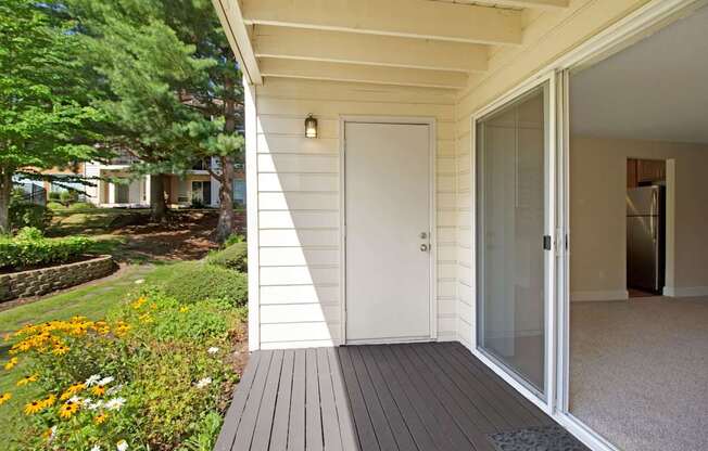 A covered private patio here at Adagio featuring painted decking, an exterior storage closet, and a sliding glass door that opens directly into the living area, while a wall-mounted light fixture provides visibility and surrounding landscaped greenery, mature trees, and flowering plants create a peaceful outdoor setting for relaxing or enjoying fresh air.