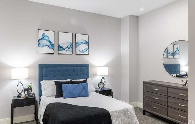 A bedroom with a large bed, a nightstand with a lamp, and a dresser with a mirror.