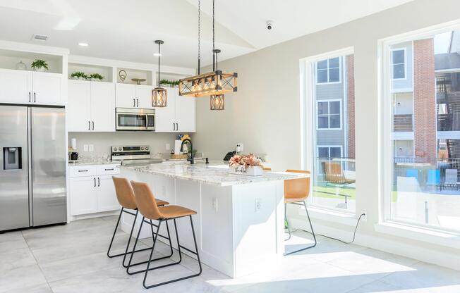 Bright modern kitchen featuring white cabinets, stainless steel appliances, and a large island with seating. A stylish chandelier hangs above the island, and large windows provide natural light, revealing an outdoor view. The space is clean and contemporary, with a neutral color palette.