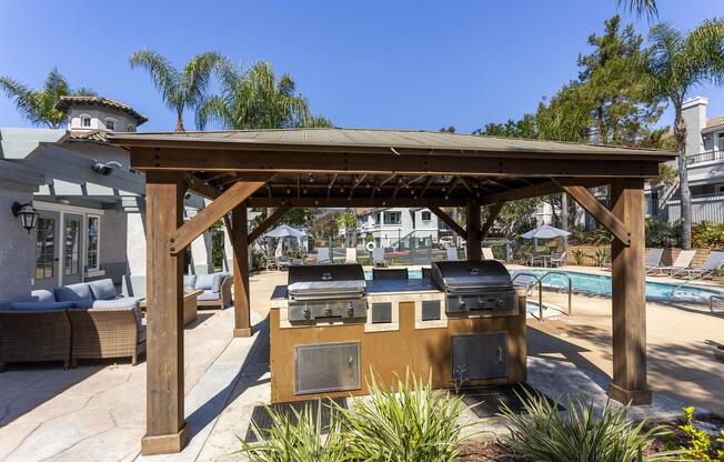 An outdoor barbecue area under a wooden gazebo, featuring two grilling stations, surrounded by palm trees and a swimming pool in the background. Comfortable seating is arranged nearby, with lounge chairs along the poolside, creating a relaxing resort-like atmosphere.