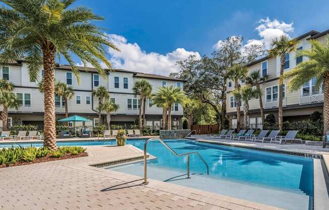 A swimming pool surrounded by palm trees and lounge chairs in front of apartment buildings.