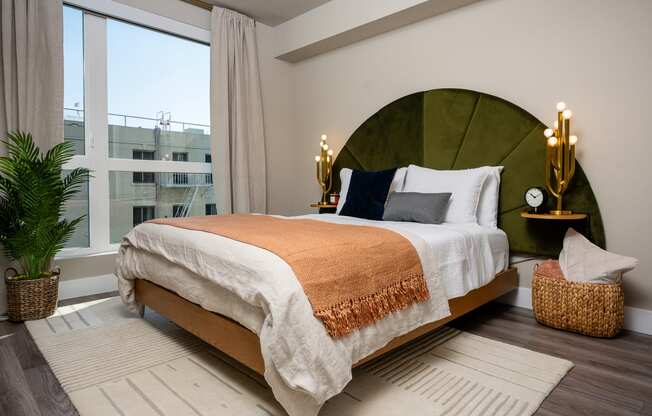 Pet-Friendly Apartments In South Park Los Angeles CA - Emerald - A Spacious And Bright Bedroom With A Full-Size Bed In The Center With One Wall Featuring A Nearly Floor-To-Ceiling Window, Wood-Style Flooring, Area Rug, And Lightly Colored Walls