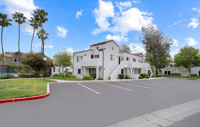 A modern multi-unit residential building surrounded by greenery, with palm trees in the background. In the foreground, there's a paved parking lot with several empty spaces. The sky is blue with a few clouds, creating a bright and inviting atmosphere.