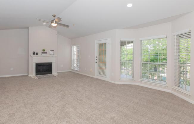 Spacious living room featuring light-colored walls, a ceiling fan, and a cozy fireplace. Large windows with white blinds allow natural light to fill the room, while plush carpet flooring adds warmth. The area is bright and inviting, with access to outdoor views.