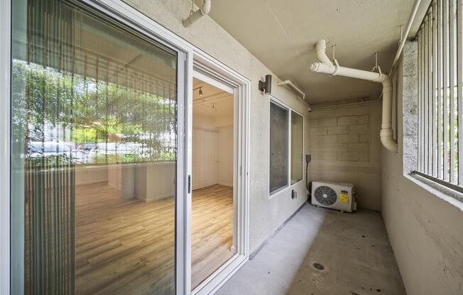 A covered patio area with sliding glass doors leading to an interior space. The floor is concrete, and there is an air conditioning unit visible. Natural light filters in, highlighting the wooden flooring inside the room.