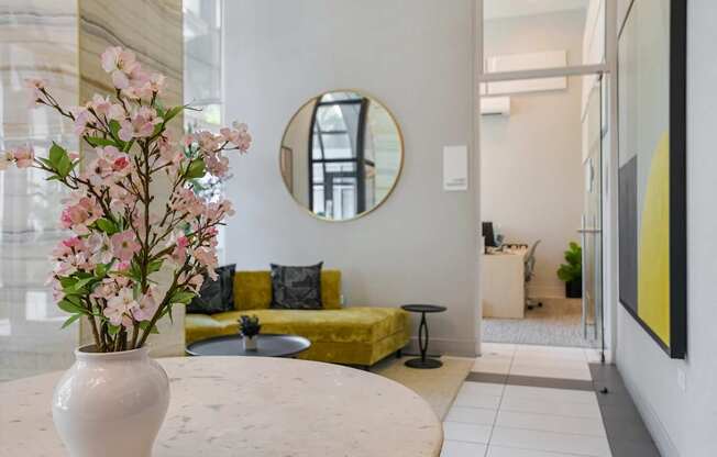 A white vase with pink flowers sits on a table in a room with a yellow couch and a round mirror on the wall.