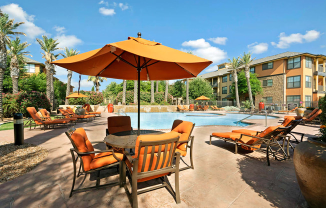 A sunny poolside gathering area here at Tuscany at Faudree featuring a resort-style swimming pool surrounded by orange cushioned lounge chairs, shaded seating with umbrellas, and a spacious tan pool deck. Palm trees, landscaped greenery, and Mediterranean-inspired apartment buildings frame the scene under a bright blue sky.