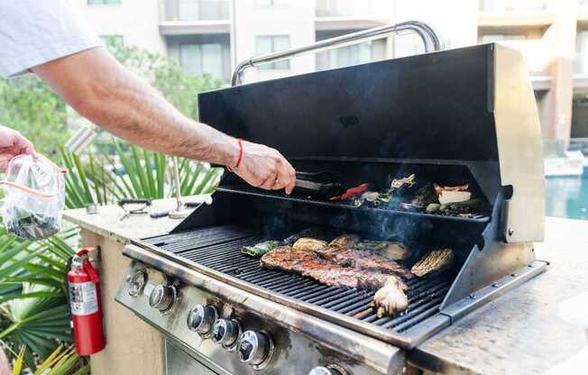 A man grilling meat on a barbecue with a red fire extinguisher nearby.