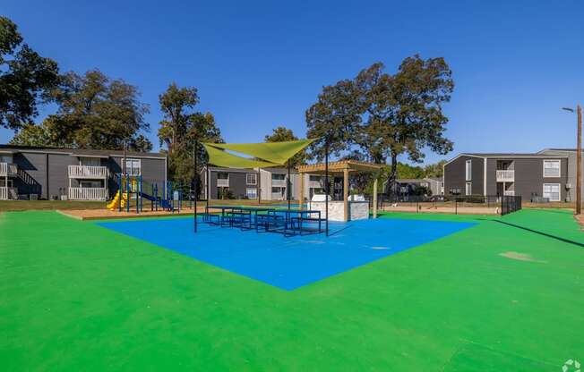 BBQ and picnic area with a blue and green surface and a yellow sunshade at Maplewood apartments in Shreveport, LA.
