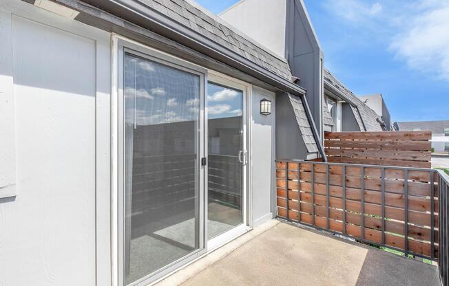A view of an outdoor balcony with sliding glass doors, surrounded by a wooden railing. The balcony features a concrete floor and is part of a residential building, with a clear blue sky in the background.