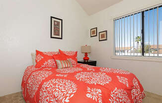 A bed with a red and white comforter is in the foreground of a room with a window.