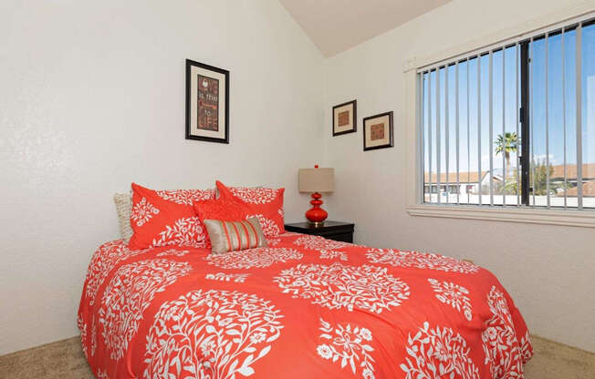 A bed with a red and white comforter is in the foreground of a room with a window.