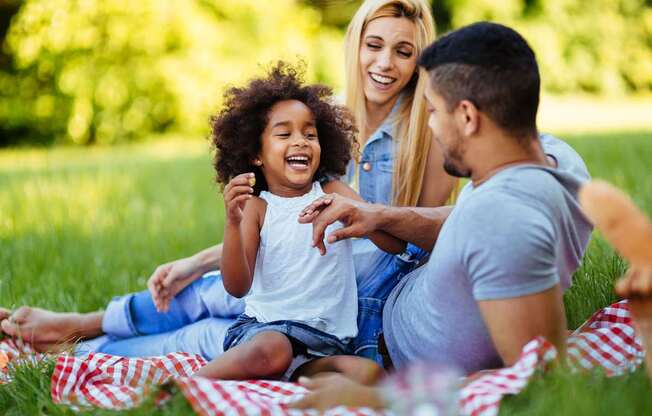 A family of three is sitting on a blanket in a park.