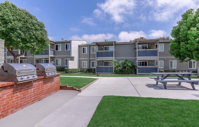 A well-maintained apartment complex with multiple buildings. In the foreground, there are barbecue grills and a picnic table on a grassy area. The background features the apartment units, which have balconies and windows, under a clear blue sky with some clouds.