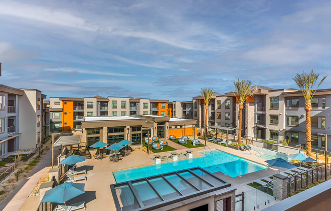 A large swimming pool is surrounded by sun loungers and umbrellas in a sunny courtyard.