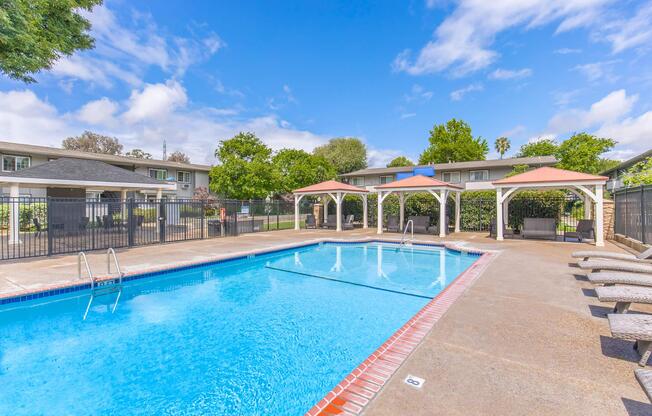 A clear blue swimming pool surrounded by a fenced area. Two shaded gazebos provide seating on one side, with lounge chairs arranged nearby. Lush greenery and trees are visible in the background under a bright blue sky with fluffy clouds. The scene suggests a relaxing outdoor space for leisure.
