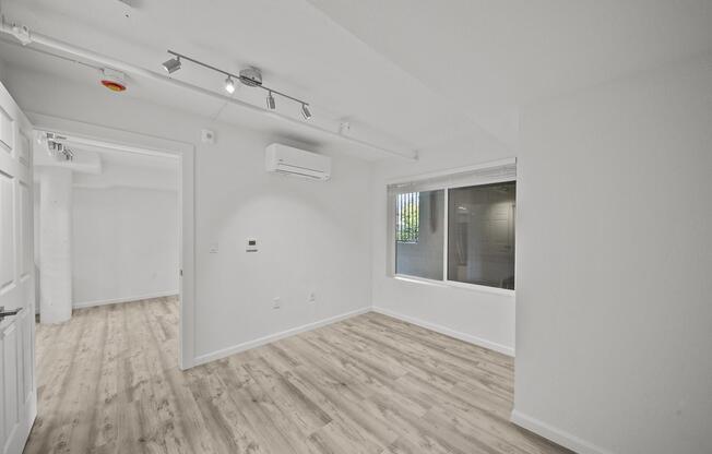 A bright, empty room with light-colored walls and wood-like flooring. There is a large window with blinds on one wall, providing natural light. An air conditioning unit is mounted near the ceiling. Another room is visible through an open door, indicating a spacious layout.