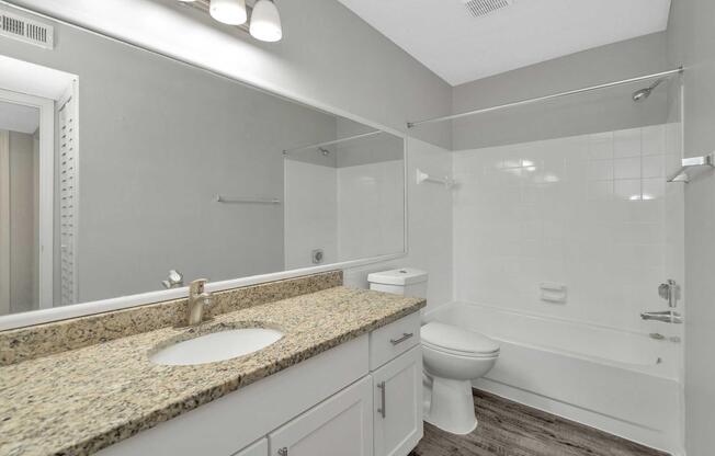 A modern bathroom featuring a granite countertop sink, a large mirror, and a white bathtub with a shower. The walls are painted in a light gray, and the flooring is a wood-like material. There is a white toilet and a towel rack in the space. Natural light is coming from a doorway.