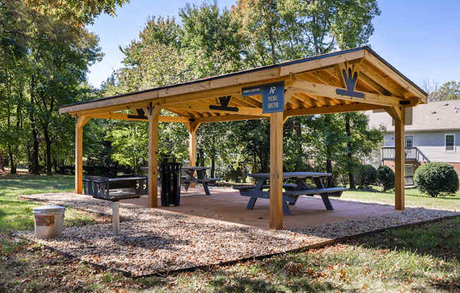 A wooden pavilion with a picnic table and benches is surrounded by trees.