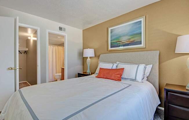 A cozy bedroom here at Northridge Court featuring a queen-size bed with crisp white bedding and accent pillows, a soft upholstered headboard, matching nightstands with lamps, warm neutral walls, framed artwork above the bed, carpeted flooring, and an open doorway leading to a private bathroom and closet area.