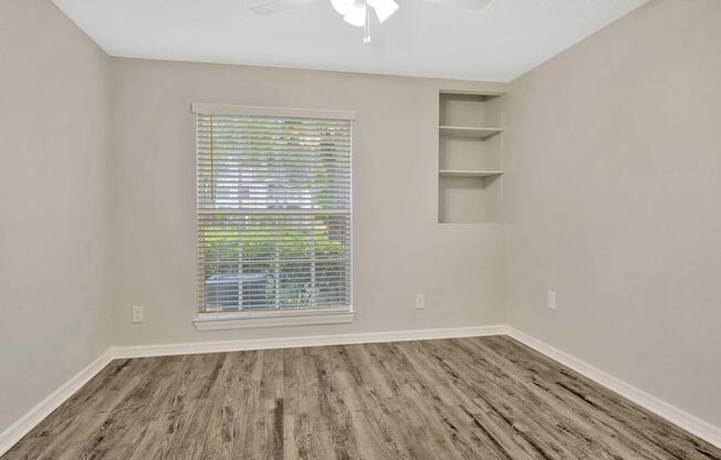 A well-lit empty room with light gray walls, featuring a window with blinds, a ceiling fan, and a wooden floor. There are built-in shelves on one wall, and the space is free of furniture, creating a clean and open atmosphere.