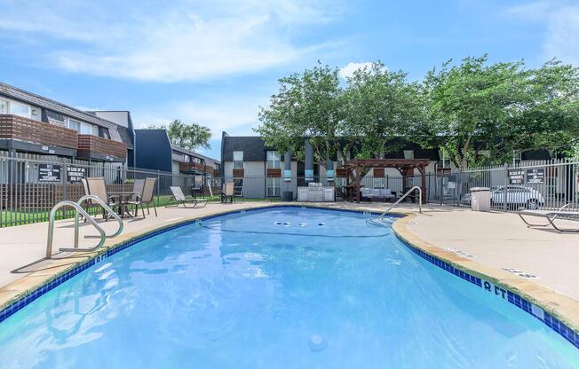 A clear blue swimming pool surrounded by a tan deck, lounge chairs, and shaded areas. Apartment buildings with balconies are visible in the background, along with trees providing some greenery. The sky is bright with a few clouds, creating a relaxed outdoor atmosphere.