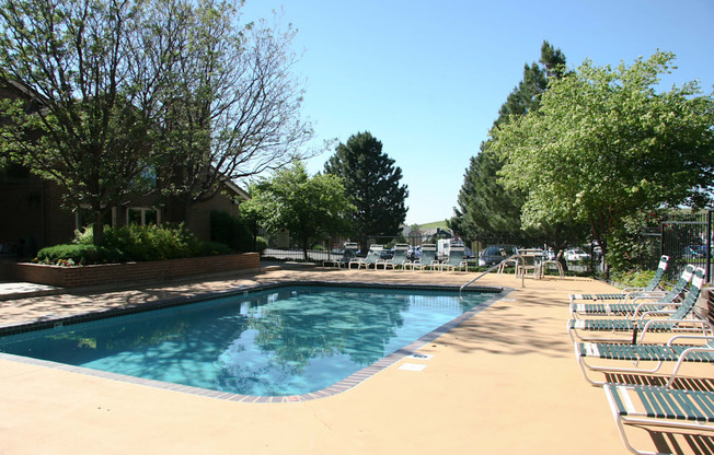 A sunny outdoor pool area here at Eagle Ridge featuring a clear rectangular swimming pool surrounded by a spacious concrete deck, neatly arranged lounge chairs, mature trees providing natural shade, and a secure fence enclosing the space, creating a relaxing and inviting community amenity on a bright day.