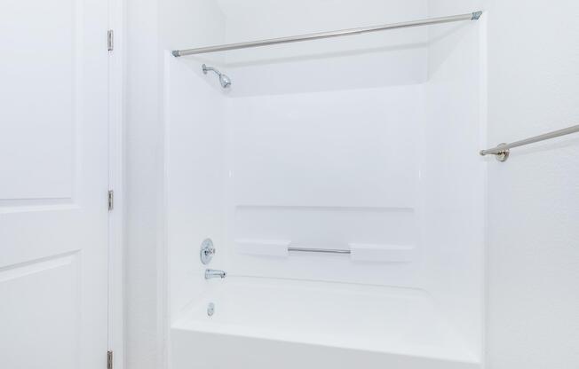 A clean, modern bathroom featuring a white tub with a shower curtain rod, minimalist design, and smooth white walls. The space appears well-lit and spacious, with a door visible in the background.