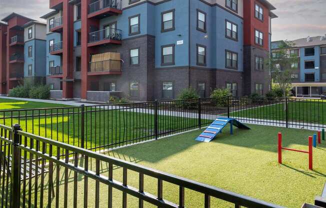A black fence surrounds a playground in front of a multi-story apartment building.